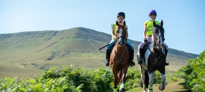Two horses and riders on narrow path with upland hillside behind in gentle sunshine