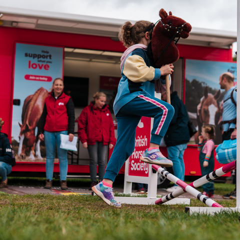 Young girl riding a hobby horse jumping a fence at a stand at a BHS event