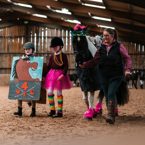 Two children in fancy dress walking alongside a horse being led by an adult in an inside arena
